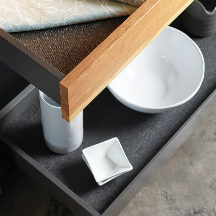 A close-up of a dark charcoal gray drawer or shelf with a light natural wood trim, containing white ceramic dishes including a bowl and small square plates, with a Hafele fiber non slip mat draped over the top edge.