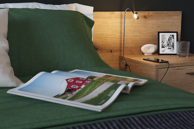 Häfele flexible LED reading light with USB charging beside a bed, providing warm task lighting over a wood headboard and nightstand.